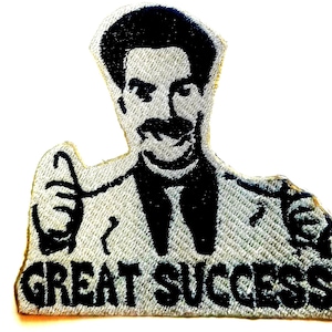 May include: Embroidered patch featuring a black and white image of a man with a mustache giving a thumbs up. The text "GREAT SUCCESS!" is below the image. The patch has a light gray background.
