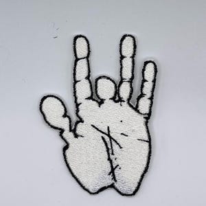 May include: White embroidered patch in the shape of a hand with the fingers in a specific gesture. The patch has a black outline and black stitching details on the palm. The background is plain white.