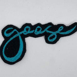 May include: Black embroidered patch with the word "goose" in teal script. The patch has a black border and a teal cursive font. The patch is likely for clothing or accessories.