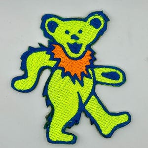 May include: Embroidered patch featuring a neon green bear with blue outlines and an orange collar. The bear is in a dancing pose with its arms and legs outstretched, and a smiling face. The patch is likely for clothing or accessories.