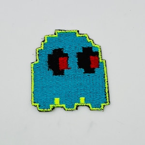 May include: A teal pixelated ghost patch with black eyes and red pupils. The ghost has a neon green outline and is likely an iron-on patch for clothing or accessories. The design is reminiscent of classic arcade games.