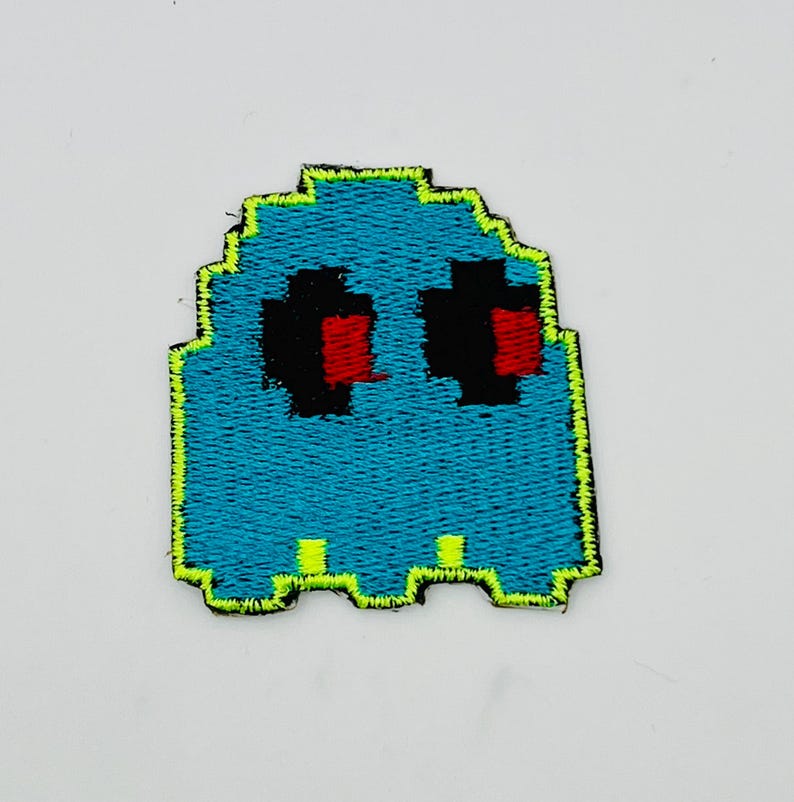 May include: A teal pixelated ghost patch with black eyes and red pupils. The ghost has a neon green outline and is likely an iron-on patch for clothing or accessories. The design is reminiscent of classic arcade games.