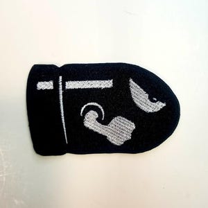 May include: Black and white embroidered patch of a video game character. The character is a black silhouette with white details. The character is shaped like a bullet.
