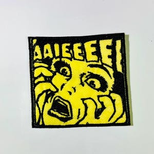 May include: Square patch with a yellow background and a black outline. The patch features a distressed illustration of a screaming face with the word "AAIEEEEE!" in black letters above the face.