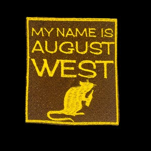 May include: A brown rectangular patch with yellow embroidered text that reads "MY NAME IS AUGUST WEST". Below the text is a yellow silhouette of a rat holding a small object. The patch is against a black background.