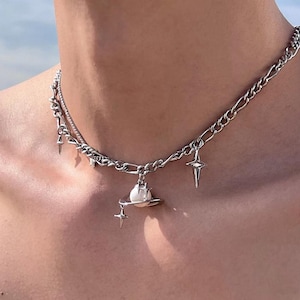 Puede incluir: Collar plateado con diseño de doble capa, compuesto por una cadena y un hilo más fino. Está adornado con colgantes en forma de estrella y un colgante que representa un planeta con una perla. La joya se muestra sobre un fondo neutro.