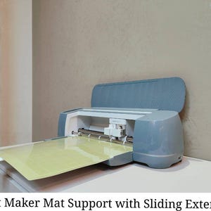 Puede incluir: Una máquina Cricut Maker azul y blanca con el soporte de la esterilla extendido. La máquina está sobre una superficie blanca con una esterilla de corte amarilla. El texto "Cricut Maker Mat Support with Sliding Extension" está en la parte inferior de la imagen.