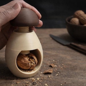 Può includere: Uno schiaccianoci in legno con la parte superiore marrone e il corpo color crema, che tiene una noce. Una ciotola di noci e un coltello sono sullo sfondo. Lo schiaccianoci è progettato per rompere le noci.