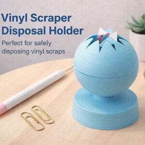 Può includere: Un supporto per lo smaltimento di raschietti in vinile azzurro con una parte superiore sferica e una base a piedistallo. Il testo "Vinyl Scraper Disposal Holder" è visibile. Un utensile artigianale bianco e rosa e graffette dorate sono sulla superficie di legno.
