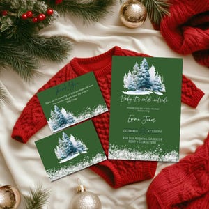 May include: A festive baby shower invitation set with a forest design. The set includes a large invitation and two smaller thank you cards. The invitations are green with a winter forest scene and the text "Baby it's cold outside". A red baby onesie is in the background.