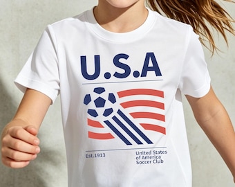 May include: White t-shirt with a graphic of the USA flag and a soccer ball. The text "U.S.A" is above the flag design, with "Est.1913" and "United States of America Soccer Club" below. The shirt is worn by a young person.