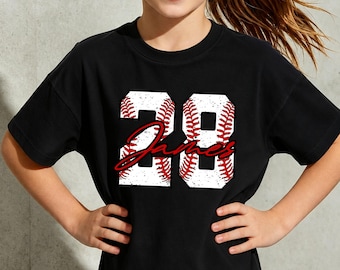 May include: Black t-shirt featuring the number 28 in a baseball design with red stitching and the name "James" in a cursive font. The shirt is worn by a person with their hands on their hips.