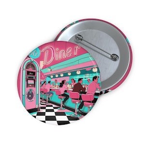 May include: A button badge with a retro diner illustration. The design features a pink and turquoise colour scheme, a jukebox, diner customers, and the word "Diner" in neon. The badge has a metal back and a pin fastening.