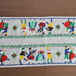 Op de afbeelding: Rechthoekige off-white tafelloper met een blauwe rand. De loper toont geborduurde figuren in traditionele klederdracht, dansend en musicerend. Groene en rode bloemaccenten zijn ook aanwezig.