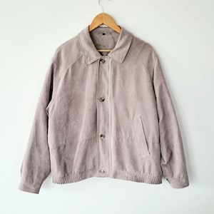 May include: A light mauve suede jacket with a collared neckline, button closure, and two front pockets. The jacket has a ribbed hem and cuffs. It is hanging on a wooden hanger against a white background.