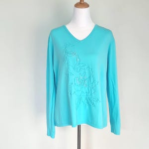 May include: A turquoise V-neck jumper with long sleeves. The jumper has an embroidered floral design on the front, with small, sparkling embellishments. It is displayed on a wooden mannequin against a plain white background.
