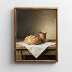 Pode incluir: Pintura emoldurada de natureza morta com um pão, uma chávena de terracota e um pano branco sobre uma mesa de madeira. O pão é castanho dourado e a chávena tem um tom quente e terroso. O fundo é de cor neutra.