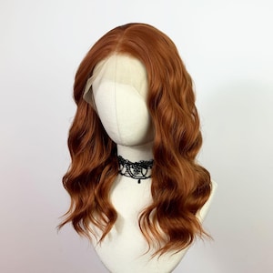 Puede incluir: Peluca ondulada de color cobre con frente de encaje, exhibida en una cabeza de maniquí. La peluca presenta mechones largos y fluidos y una línea de cabello de aspecto natural. Se usa un collar de gargantilla de encaje negro.