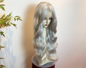 May include: A long, wavy wig in a light silver-gray color with a center part. The hair features soft, voluminous curls that cascade down past the shoulders, displayed on a mannequin head with a visible lace front hairline.