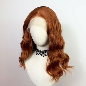 May include: A copper-colored, wavy wig with a lace front is displayed on a mannequin head. The wig has a natural-looking hairline and is styled with loose waves. A black lace choker necklace is around the mannequin's neck.