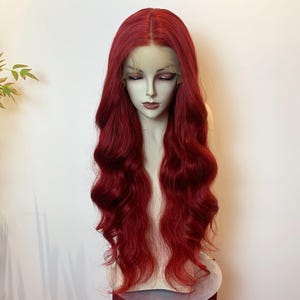 Puede incluir: Una larga peluca roja ondulada sobre una cabeza de maniquí. La peluca presenta un frente de encaje y un color rojo intenso y rico. El cabello cae en ondas voluminosas, creando un aspecto dramático y elegante. El maniquí tiene una tez pálida.