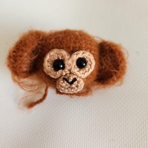 May include: A crocheted monkey head with fuzzy rust-colored yarn, large round ears, and a light tan face patch. It features two shiny black plastic safety eyes and a small embroidered black nose and mouth detail on the muzzle area.