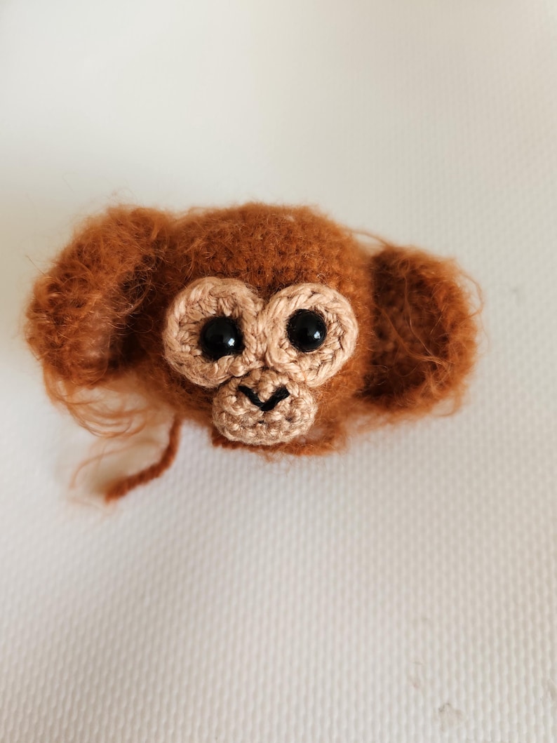 May include: A crocheted monkey head with fuzzy rust-colored yarn, large round ears, and a light tan face patch. It features two shiny black plastic safety eyes and a small embroidered black nose and mouth detail on the muzzle area.