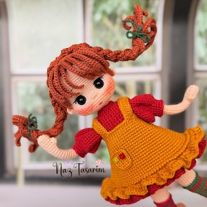 May include: A handcrafted crochet doll with vibrant orange yarn hair styled in two braided pigtails tied with green ribbons. The doll wears a red short-sleeved dress under a mustard yellow pinafore with a small pocket and text Naz Tasarım.