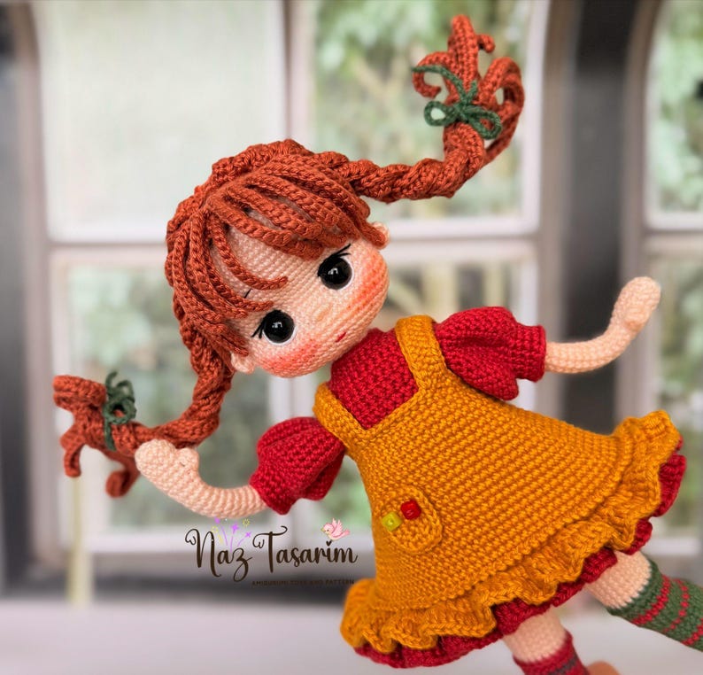 May include: A handcrafted crochet doll with vibrant orange yarn hair styled in two braided pigtails tied with green ribbons. The doll wears a red short-sleeved dress under a mustard yellow pinafore with a small pocket and text Naz Tasarım.