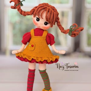 May include: A crocheted doll with auburn hair in two braids, wearing a red short-sleeved shirt under a mustard yellow pinafore dress with a ruffled hem. The doll has striped socks in red, green, and brown, and yellow shoes. Text reads Naz Tasarım.