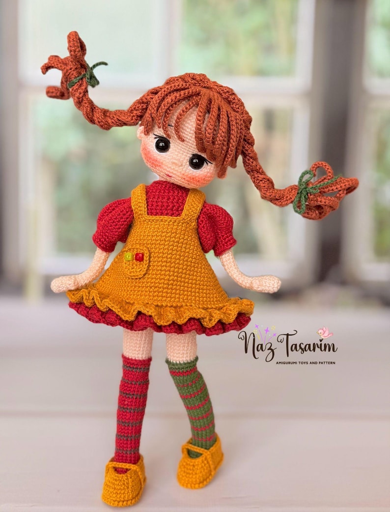May include: A crocheted doll with auburn hair in two braids, wearing a red short-sleeved shirt under a mustard yellow pinafore dress with a ruffled hem. The doll has striped socks in red, green, and brown, and yellow shoes. Text reads Naz Tasarım.