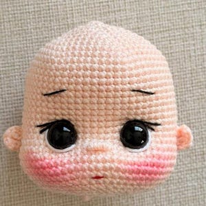 May include: A crocheted doll head made with light peach yarn, featuring large black plastic eyes, embroidered black eyebrows and eyelashes, a small red mouth, and pink blush accents on the cheeks. The head has two small ears on the sides.