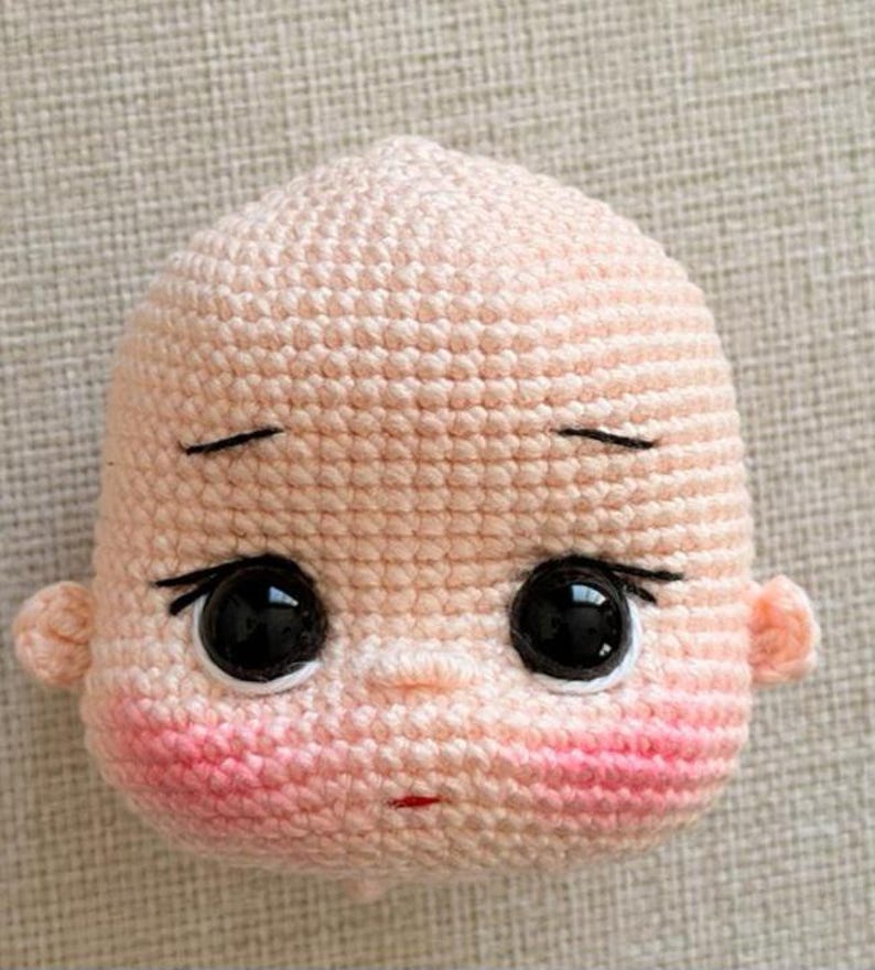 May include: A crocheted doll head made with light peach yarn, featuring large black plastic eyes, embroidered black eyebrows and eyelashes, a small red mouth, and pink blush accents on the cheeks. The head has two small ears on the sides.