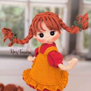 May include: A handcrafted crochet doll with vibrant orange yarn hair styled in two braided pigtails tied with green bows. The doll wears a mustard yellow pinafore dress over a red short-sleeved shirt. The text Naz Tasarım is visible on the left.
