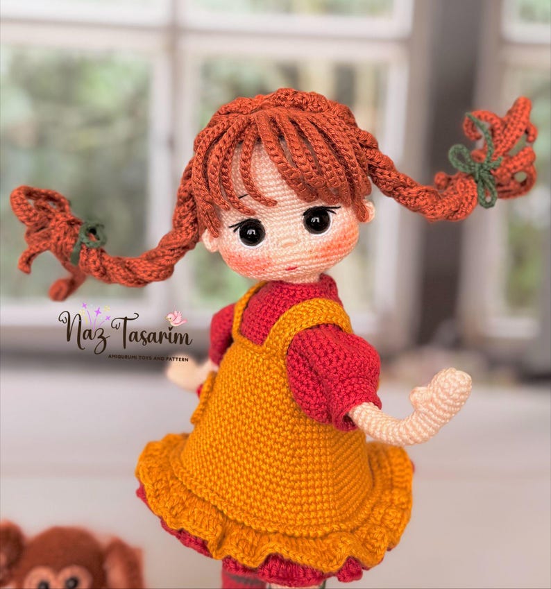 May include: A handcrafted crochet doll with vibrant orange yarn hair styled in two braided pigtails tied with green bows. The doll wears a mustard yellow pinafore dress over a red short-sleeved shirt. The text Naz Tasarım is visible on the left.