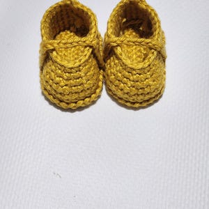 May include: A pair of mustard yellow baby booties crafted with a textured crochet stitch. Each shoe features a rounded toe, a structured ankle opening, and a decorative strap detail across the top, showcasing a consistent yarn construction.