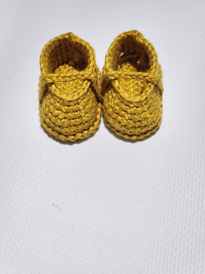 May include: A pair of mustard yellow baby booties crafted with a textured crochet stitch. Each shoe features a rounded toe, a structured ankle opening, and a decorative strap detail across the top, showcasing a consistent yarn construction.