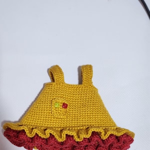 May include: A mustard yellow crocheted dress with two shoulder straps, a small chest pocket featuring two tiny square beads in lime green and red, and a ruffled hemline in a deep maroon color, crafted with a dense, textured yarn stitch pattern.