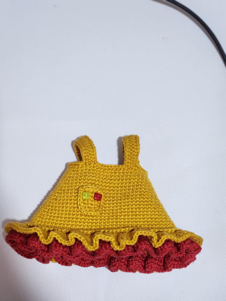 May include: A mustard yellow crocheted dress with two shoulder straps, a small chest pocket featuring two tiny square beads in lime green and red, and a ruffled hemline in a deep maroon color, crafted with a dense, textured yarn stitch pattern.