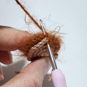 May include: A metal crochet hook with a light pink handle is being used to work with fuzzy, rust-colored yarn. The hook is inserted into a small, circular, tan-colored crocheted piece, demonstrating the process of attaching textured yarn.