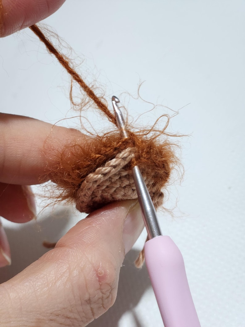 May include: A metal crochet hook with a light pink handle is being used to work with fuzzy, rust-colored yarn. The hook is inserted into a small, circular, tan-colored crocheted piece, demonstrating the process of attaching textured yarn.