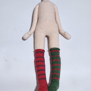 May include: A crochet doll body with a light beige torso and arms. The doll features mismatched legs: the left leg has red and dark blue horizontal stripes, while the right leg has green and red horizontal stripes. The doll has no head.