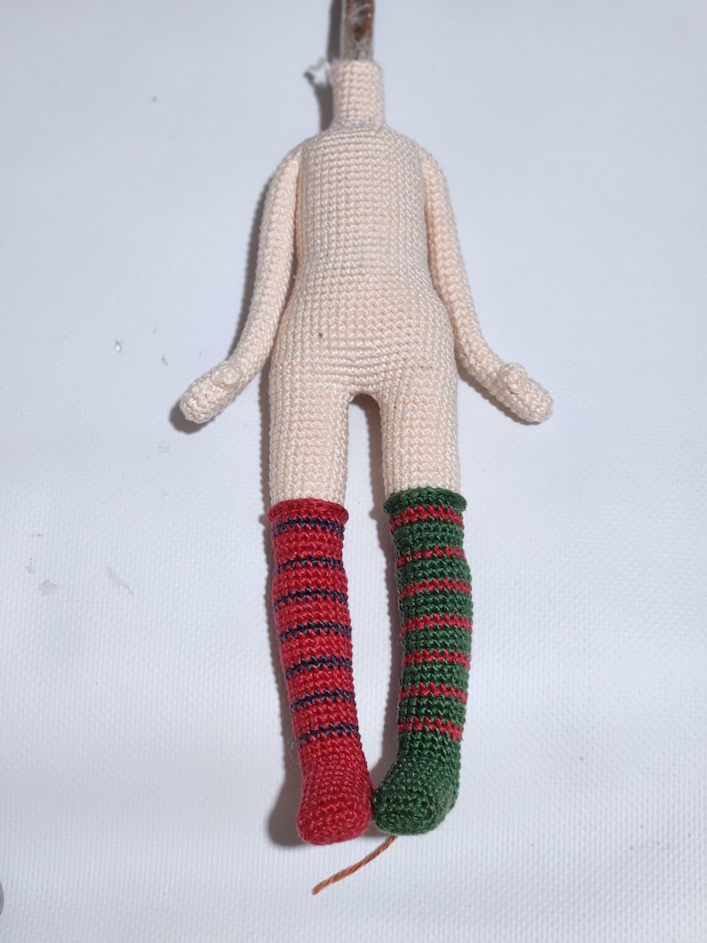 May include: A crochet doll body with a light beige torso and arms. The doll features mismatched legs: the left leg has red and dark blue horizontal stripes, while the right leg has green and red horizontal stripes. The doll has no head.