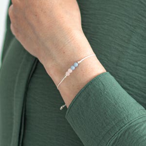 Puede incluir: Pulsera delicada con tres cuentas de colores pastel: rosa, azul claro y rosa más claro. Las cuentas están ensartadas en un cordón fino plateado. La pulsera se muestra en una muñeca, sobre un fondo de camisa verde.