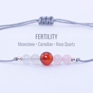 May include: A gray adjustable bracelet with the word "FERTILITY" and the names of the stones: Moonstone, Carnelian, and Rose Quartz. The bracelet features a series of round beads in white, red, and pink.