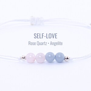 May include: A white cord bracelet with pink rose quartz and blue angelite beads, and silver details. The text "SELF-LOVE" is above the words "Rose Quartz • Angelite".