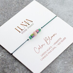 Puede incluir: Una delicada pulsera con cuentas verdes, rosas e iridiscentes ensartadas en un cordón verde oscuro. La pulsera se muestra en una tarjeta blanca con las palabras "Calm Bloom" y "LUMA STONE POWER" impresas.