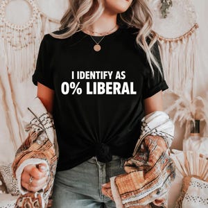 May include: Black t-shirt with the white text "I IDENTIFY AS 0% LIBERAL." The shirt is knotted at the waist. A patterned jacket is draped over the shoulders. The person is wearing blue jeans.