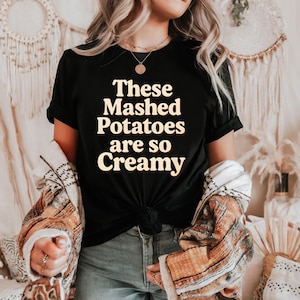 Puede incluir: Camiseta negra con el texto "These Mashed Potatoes are so Creamy" en crema. La camiseta está anudada a la cintura. La persona lleva una chaqueta estampada sobre la camiseta.