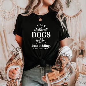 Puede incluir: Camiseta negra con el texto "A Day Without DOGS is like.... Just kidding. I HAVE NO IDEA." La camiseta está anudada a la cintura. La persona lleva una chaqueta estampada y vaqueros azules.